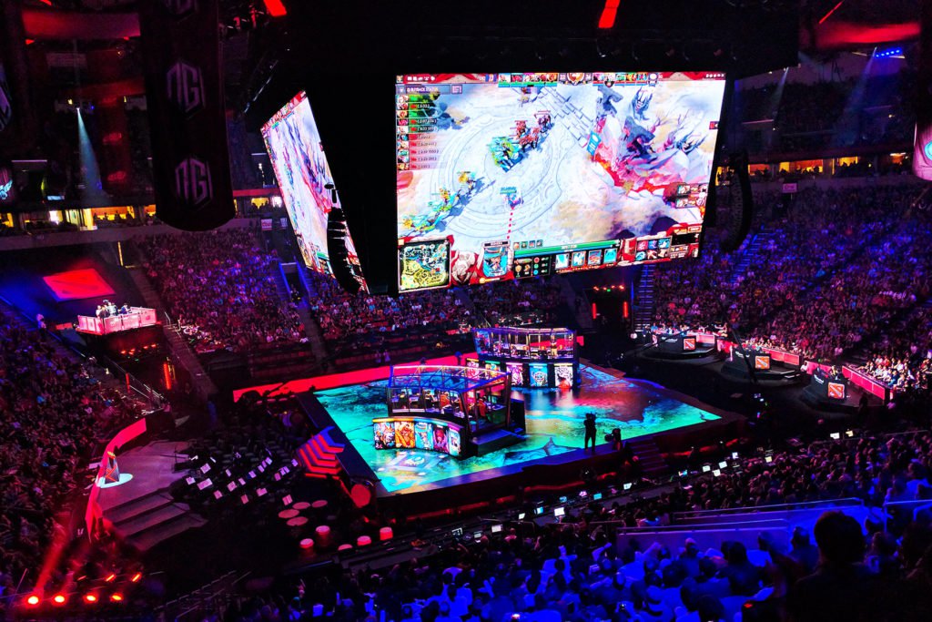 Publiek kijkt naar een esporttoernooi in een grote arena met spelers in actie op het podium en het spel op schermen erboven.